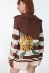 Cardigan tejido de punto color marrón con rayas horizontales blancas y bordado de flores amarillas. Tiene cuello marinero, cierre con botones de madera y corte holgado.