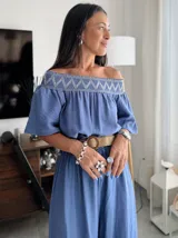 Enterito largo color blanco, con escote Bardot fruncido y mangas cortas abullonadas. La cintura es ajustable con un cinturón ancho de rafia con hebilla redonda. El pantalón es ancho y largo.