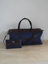 Bolso de lona azul marino con detalles en cuero marrón, doble asa de mano y correa bandolera ajustable. Incluye un neceser haciendo juego con logo de caballo plateado.