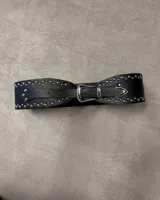 Cinturón ancho de cuero negro con doble hilera de tachas plateadas. Hebilla metálica plateada de estilo occidental y punta de cierre en forma de flecha.