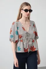 Blusa con escote en V y lazos regulables, mangas cortas con volumen y frunce bajo el busto. Presenta un estampado de flores en tonos naranja y turquesa sobre un fondo de rayas verticales blancas y negras.