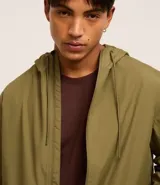 Campera rompevientos color verde oliva, con capucha ajustable con cordón, cierre frontal con cremallera, bolsillos laterales con cremallera y puños elásticos.