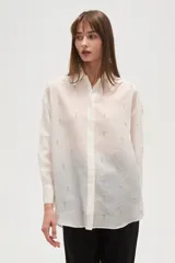 Camisa blanca de poliéster, de corte ancho y manga larga, con cuello redondo y apliques de strass y tachas distribuidos por toda la prenda.