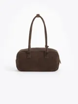 Cartera rectangular de estructura firme confeccionada en cuero descarne vacuno color marrón. Presenta manijas fijas, cierre superior y un charm metálico dorado en forma de vaquita de San Antonio colgando de un tag lateral. Incluye el logo de la marca grabado en relieve en el frente.