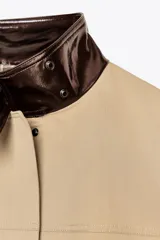 Cazadora color beige con cuello alto en tejido efecto piel marrón a contraste. Manga larga, bolsillos delanteros, detalle de pespuntes a tono, forro interior y cierre frontal con cremallera y botones a presión ocultos por solapa.