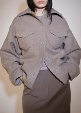 Chaqueta color gris topo de tejido bouclé, con cuello camisero, cierre frontal con botones, dos bolsillos de parche con solapa y botón en el pecho, y mangas largas.