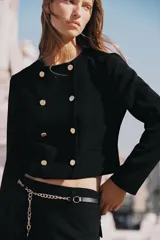 Blazer corta negra de cuello redondo y manga larga, con cierre frontal cruzado con botones dorados y falsos bolsillos delanteros.