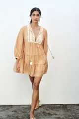 Vestido corto color beige con cuello redondo, cordón con borlas y manga larga. Presenta bordados combinados a contraste en la pechera, cierre frontal con botones ocultos, forro interior y bajo con volantes.