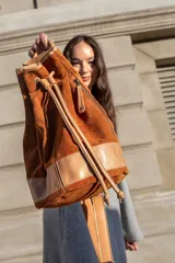 Mochila tipo bucket color marrón tostado, confeccionada en gamuza con detalles en cuero color beige. Cuenta con bolsillo exterior con cierre, bolsillo interior con cierre, llavero interno, asa de mano y correa extraíble y regulable para usar como mochila o bolso de hombro.
