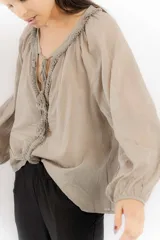 Blusa color beige con escote en V y mangas largas abullonadas. Presenta detalle de cordones trenzados con borlas en el escote.