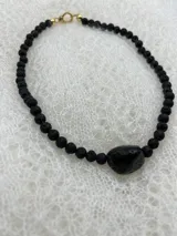 Collar corto de cuentas redondas de piedra volcánica negra, con cuenta central irregular de piedra negra pulida y cierre de gancho dorado.