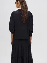 Camisa de broderie de algodón color negro, con cuello mao y cierre de cuatro botones. Presenta mangas abullonadas y galones con transparencias en la parte delantera y mangas.