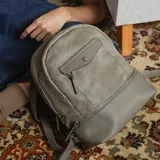 Mochila gris de símil cuero con bolsillo frontal con cierre y broche. Tiene apertura superior, cierres con tiradores en el mismo tono, bolsillos internos, correas regulables con hebilla y asa de mano.
