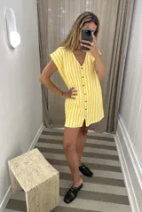 Vestido corto de tejido de punto con diseño de rayas verticales en beige y blanco. Presenta una silueta holgada, escote en V y cierre frontal con botones metálicos. Confeccionado en una mezcla de algodón y poliéster con textura de jogging sin friza, ideal para usar como salida de baño.