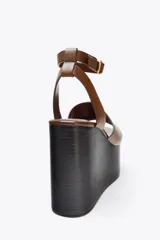 Sandalias de cuero color marrón con plataforma de cuña de madera oscura y correa de tobillo ajustable con hebilla dorada.