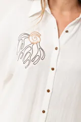 Camisa blanca de silueta oversize, con cuello camisero, manga 3/4 y corte más largo en la espalda. Presenta bordados de coral y caracol en el frente, y botones a tono.