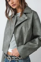 Campera biker color verde grisáceo con efecto desgastado, cuello con solapa y cierre lateral. Presenta herrajes plateados y detalles de cordones entrelazados en los codos y en la espalda.