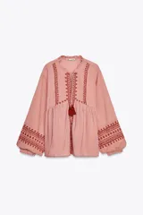 Camisa de algodón color rosa con rayas verticales finas en rojo. Tiene cuello redondo con volantes, escote pico, mangas largas con puños y bordados combinados a contraste en rojo. Cierre frontal con botones y lazada con pompones.