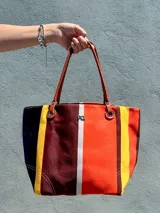 Bolso tipo tote bag de lona con diseño de rayas verticales en tonos cálidos: naranja, amarillo, marrón oscuro y blanco. Posee asas de cuero marrón y un dije metálico con las iniciales 'PQ' en el centro.