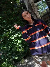 Sweater de manga larga estilo rugby, color beige, con franjas horizontales negras y rosadas. Posee cuello polo con solapa negra y un parche bordado en el pecho.