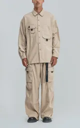 Camisa de manga larga color beige, estilo técnico con calce oversize. Presenta cuello camisero, cierre frontal de botones y múltiples bolsillos de parche con solapa en el frente, algunos con tiradores negros.