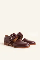 Zapato tipo monk strap con aberturas laterales, confeccionado en cuero vacuno color bordó. Presenta doble tira con hebillas metálicas plateadas en el empeine y suela de cuero con taco bajo.