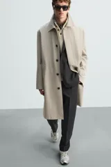 Gabardina color arena de corte recto, con cuello solapa, cierre frontal con botones, mangas largas con puños abotonados, bolsillos laterales y abertura en la parte posterior.