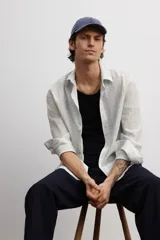 Camisa blanca de popelina de algodón texturizado, con cuello inglés, tapeta lisa, canesú con pliegue de caja en la espalda y mangas largas con abertura y cierre ajustable de botón en los puños. Corte holgado y bajo levemente redondeado.
