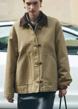 Chaqueta color beige con cuello camisero de pana marrón, cierre frontal con broches metálicos y dos bolsillos de parche delanteros.