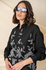 Camisa blanca de algodón con bordado floral en hilo negro en el frente. Camisa de corte clásico, con cuello abotonado, mangas largas con puños abotonados y cierre frontal con botones.