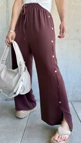 Pantalón ancho color chocolate con cintura elastizada y cordón ajustable. Presenta botones decorativos en los laterales y aberturas en los tobillos.