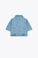 Camisa de denim celeste de corte crop, con cuello solapa, escote en pico, manga corta y dos bolsillos delanteros con botón. Cierre frontal con botones metálicos.