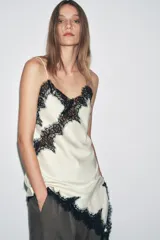 Vestido corto de satén color blanco, con tirantes finos, escote en pico y bajo asimétrico con abertura. Detalle de encaje negro en escote y ruedo.