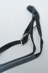 Cartera pequeña tipo clutch o bandolera de color negro, con borde decorado con tachas cuadradas plateadas. Posee cierre superior con cremallera y correa larga ajustable para llevar al hombro o cruzada.