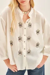 Camisa blanca de voilé de algodón con escote redondo y bordados de ojos en el delantero. De corte recto y mangas largas con puños abotonados.