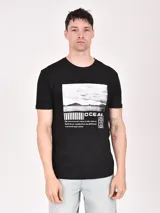 Remera negra de manga corta con estampa rectangular en el frente que incluye una foto de un paisaje marítimo, barras horizontales, la palabra "OCEAN" y un texto descriptivo.