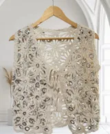Chaleco de crochet color beige, sin mangas, con diseño calado de flores y adornado con lentejuelas plateadas. Presenta un escote en V y se ata al frente con un lazo.