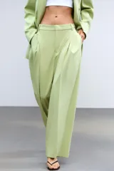 Pantalón verde claro de tiro alto con trabillas y botón en la cintura. Tiene pliegues, bolsillos laterales y falsos bolsillos de vivo en la espalda. El cierre frontal es con cremallera, botón interior y ganchos metálicos.