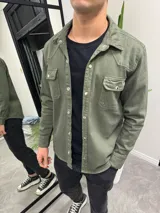 Sobrecamisa verde militar de corte recto, con cuello clásico, cierre frontal con botones metálicos, dos bolsillos con solapa en el pecho y mangas largas con puños abotonados.