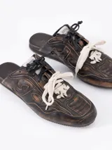 Zapatilla tipo mule (sin talón) de cuero vacuno, color celeste con efecto gastado y detalles de recortes. Presenta un logo estilizado en el lateral y cordones anchos de color beige y marrón.