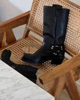 Bota alta de cuero vacuno color negro, con caña recta, puntera cuadrada y detalle de arnés con hebilla metálica plateada.