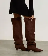 Bota over the knee de gamuza sintética color marrón, con efecto slouchy y caída fruncida a lo largo de la caña. Presenta punta fina y tacón bajo y fino de 5 cm.