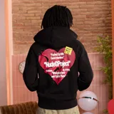 Campera negra con capucha, corte oversize y estampado de corazón roto con texto en la espalda. Incluye un sticker amarillo con texto manuscrito.