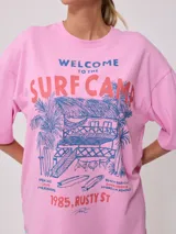 Remera blanca de algodón con estampado frontal de estilo playero con texto "Welcome to the Surf Camp" y dibujo de una casa de playa con palmeras y tablas de surf.