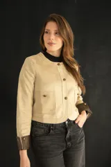 Chaqueta corta color beige con botones dorados, cuello redondo y puños marrones en contraste.
