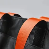 Bolso tote negro de tela resistente al agua, con asas de correa negras con borde naranja. Tiene cierre principal, bolsillos laterales (uno con cierre), seis bolsillos internos, diseño plegable y capacidad de 14,5 litros.