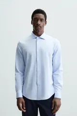 Camisa celeste a rayas verticales blancas, de corte recto, con cuello italiano abierto, mangas largas con puños abotonados y cierre frontal con botones.