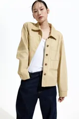Campera color beige de sarga de algodón, con cuello, botones al frente y canesú en la espalda. Tiene hombros caídos, bolsillos superiores de parche, bolsillos insertados al frente y puños abotonados.