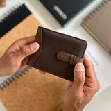 Billetera marrón oscuro de símil cuero y tela canvas con presilla. Interior con visor, doble repartición para billetes, tarjeteros y bolsillo.
