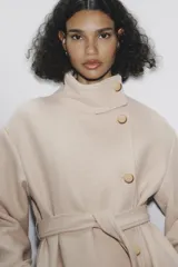 Abrigo largo color beige confeccionado en hilatura con lana. Tiene cuello subido, manga larga con trabillas, bolsillos laterales y cinturón ajustable con lazada. El cierre frontal es cruzado con botones.
